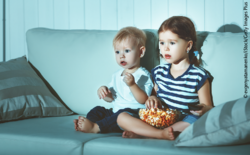 Zwei Kleinkinder sitzen mit einer Schüssel Popcorn auf dem Sofa.